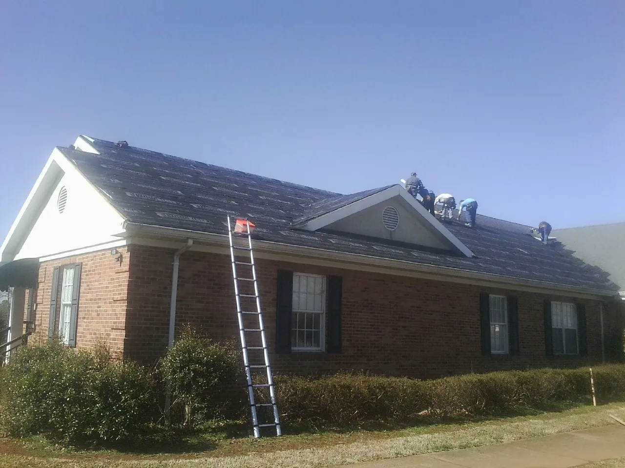 Crew tearing shingles off a brick ranch roof with a ladder set — Emergency Roof Repair work by our crew in Reydon