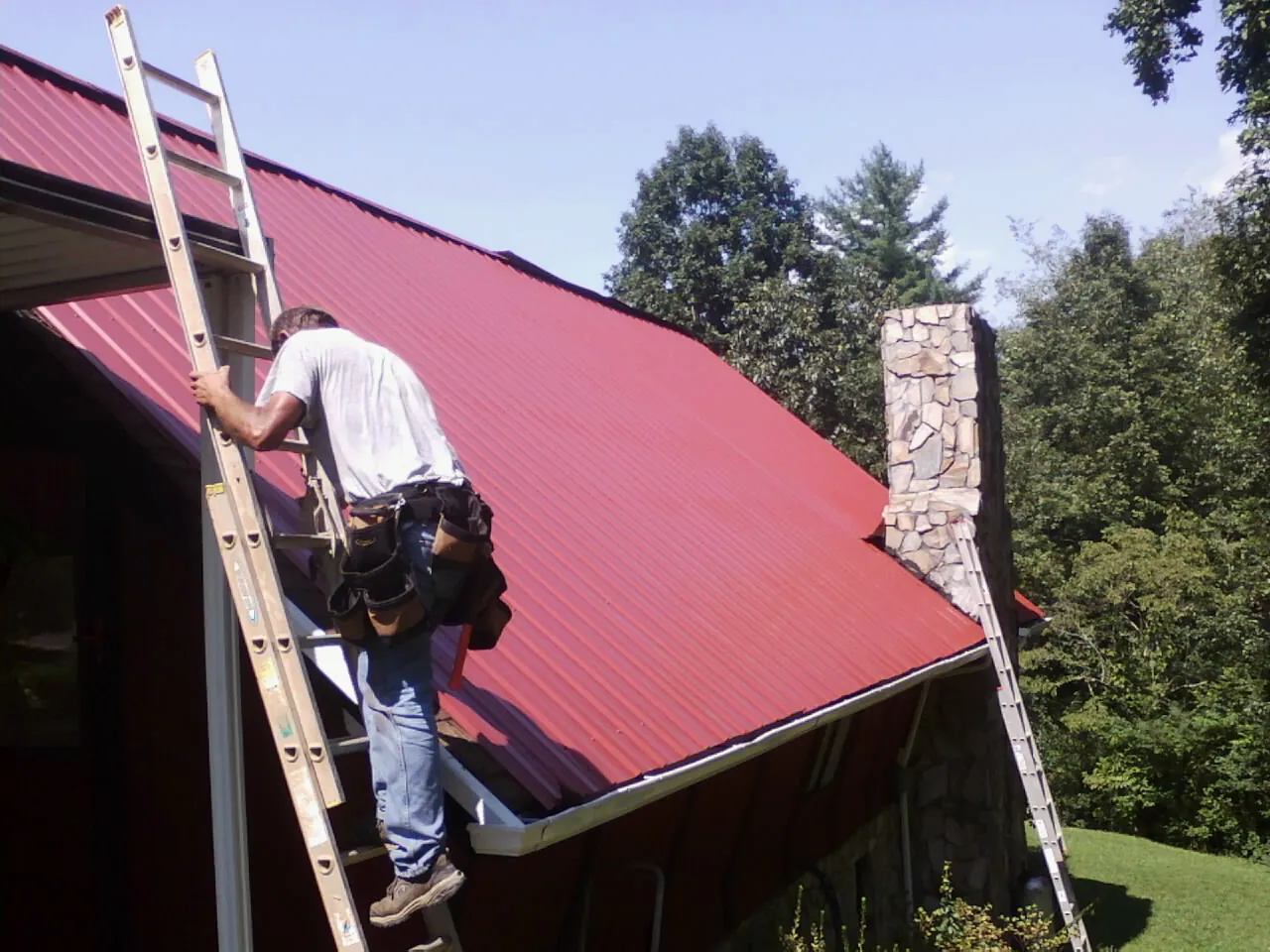 Roofer on a ladder beside a red metal roof and stone chimney — Soffit & Fascia Repair by Roofing Brothers in Reydon